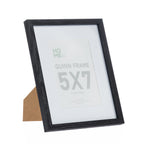 A black woodgrain 5×7-inch photo frame with a white mat is displayed on its easel stand against a plain light background.