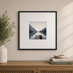 A black woodgrain square photo frame with a white mat showcases a tranquil lakeside landscape and is styled above a wooden console with neutral décor in a contemporary interior.