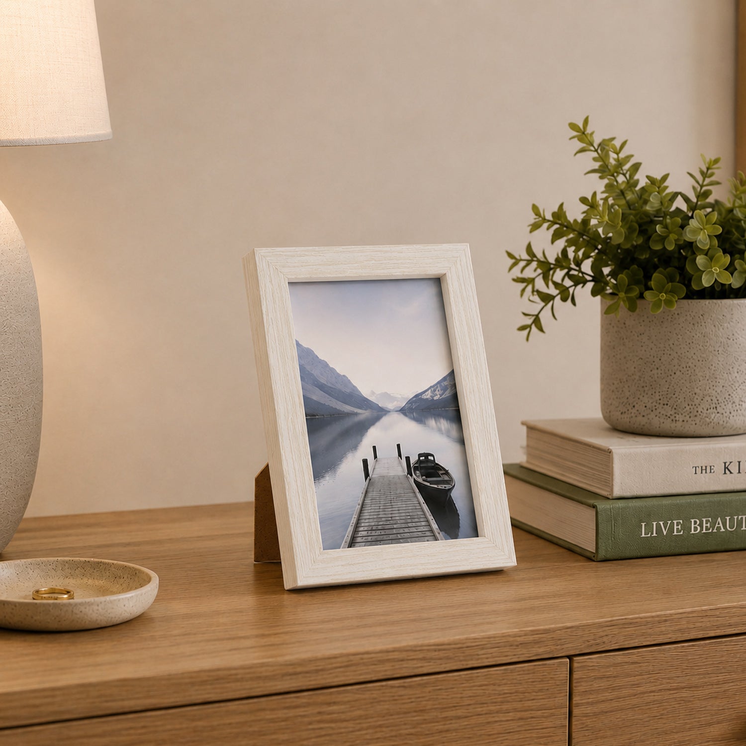 A white wood-look photo frame displaying a tranquil lakeside landscape is styled on a timber console alongside books, greenery, and a ceramic lamp for a warm, contemporary look.