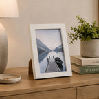 A white wood-look photo frame displaying a tranquil lakeside scene adds a timeless decorative touch to a timber console styled with books, greenery, and a ceramic lamp.