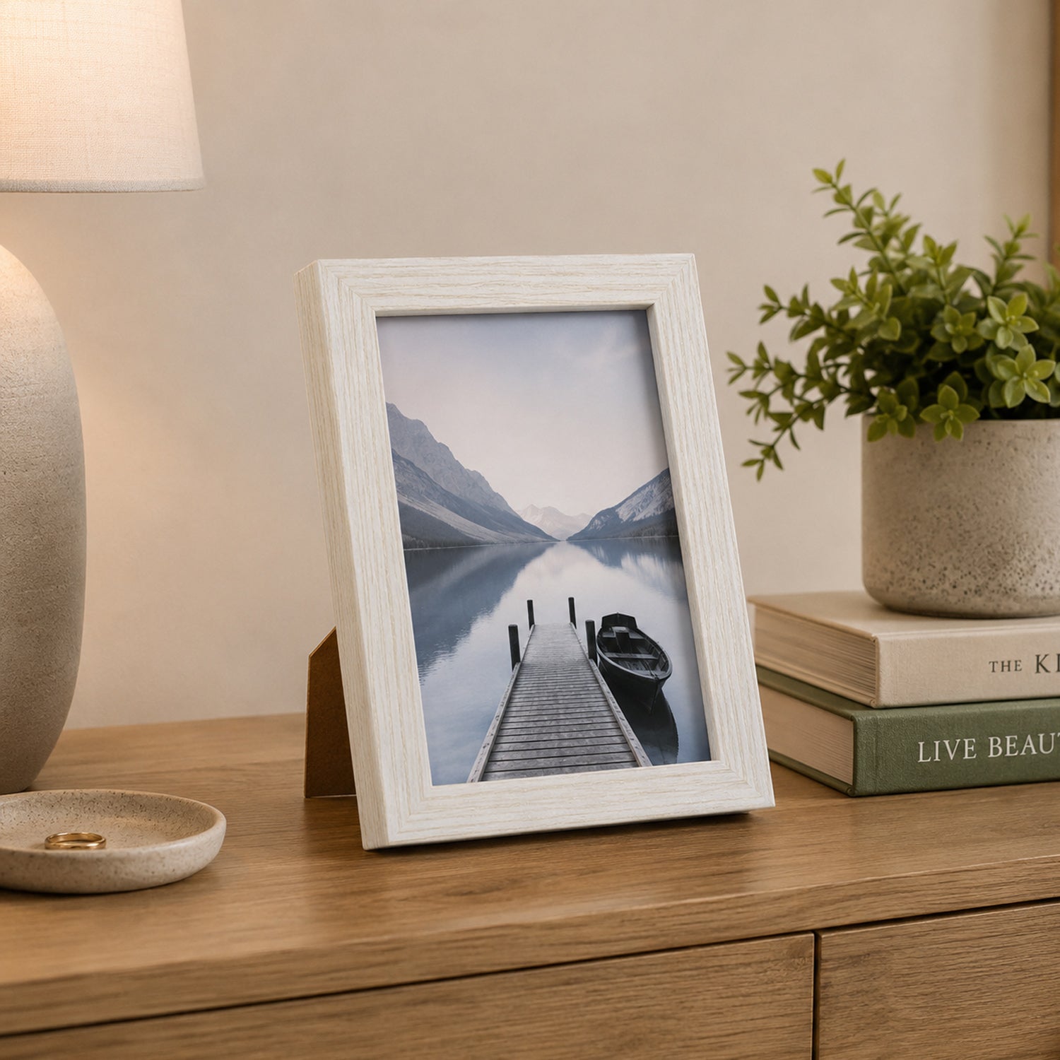 A white wood-look photo frame displaying a tranquil lakeside scene adds a timeless decorative touch to a timber console styled with books, greenery, and a ceramic lamp.