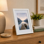 A white wood-look photo frame displaying a tranquil lakeside landscape is styled on a timber console alongside books, greenery, and a ceramic lamp for a warm, contemporary look.