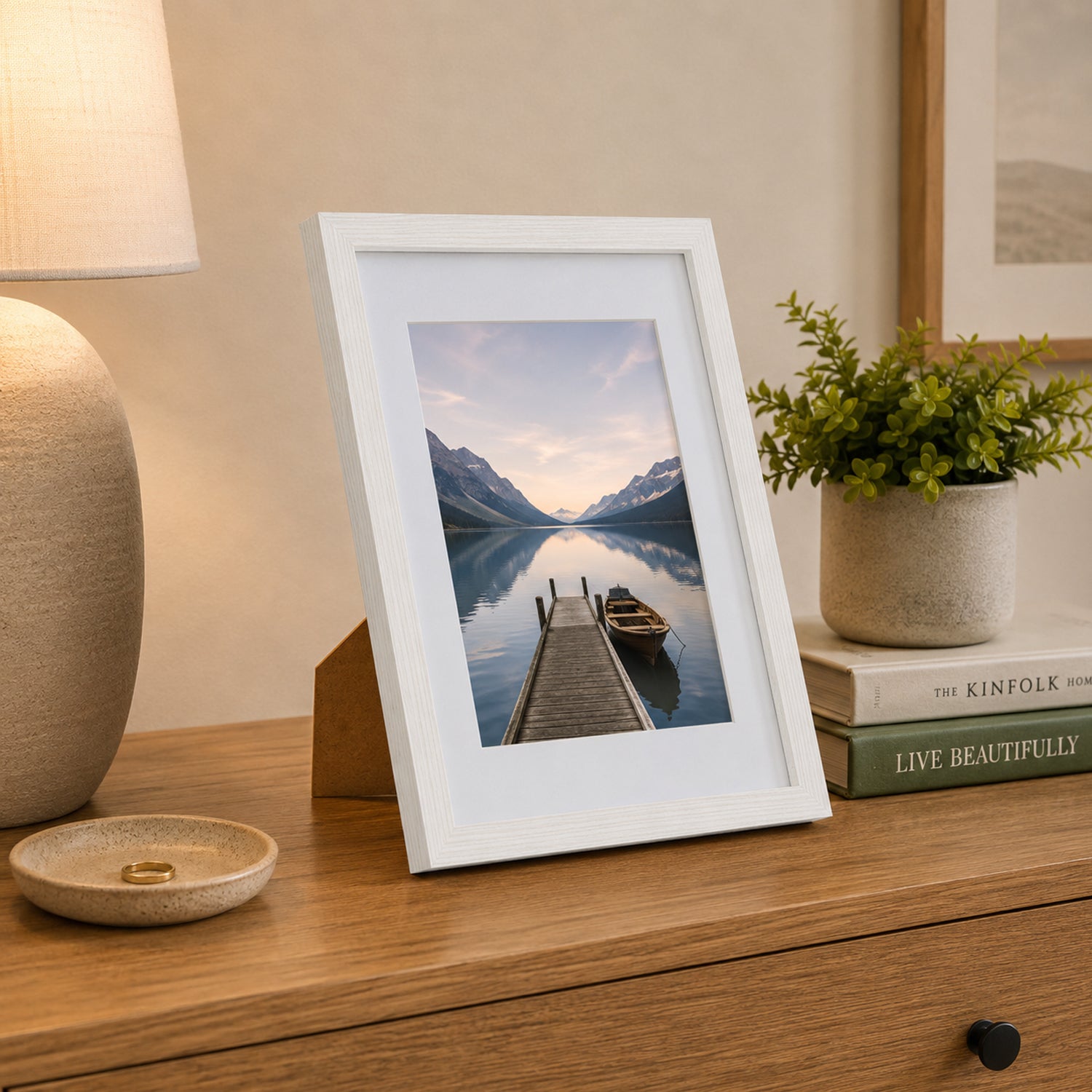 A white wood-look photo frame displaying a tranquil lakeside landscape is styled on a timber console alongside books, greenery, and a ceramic lamp for a warm, contemporary look.