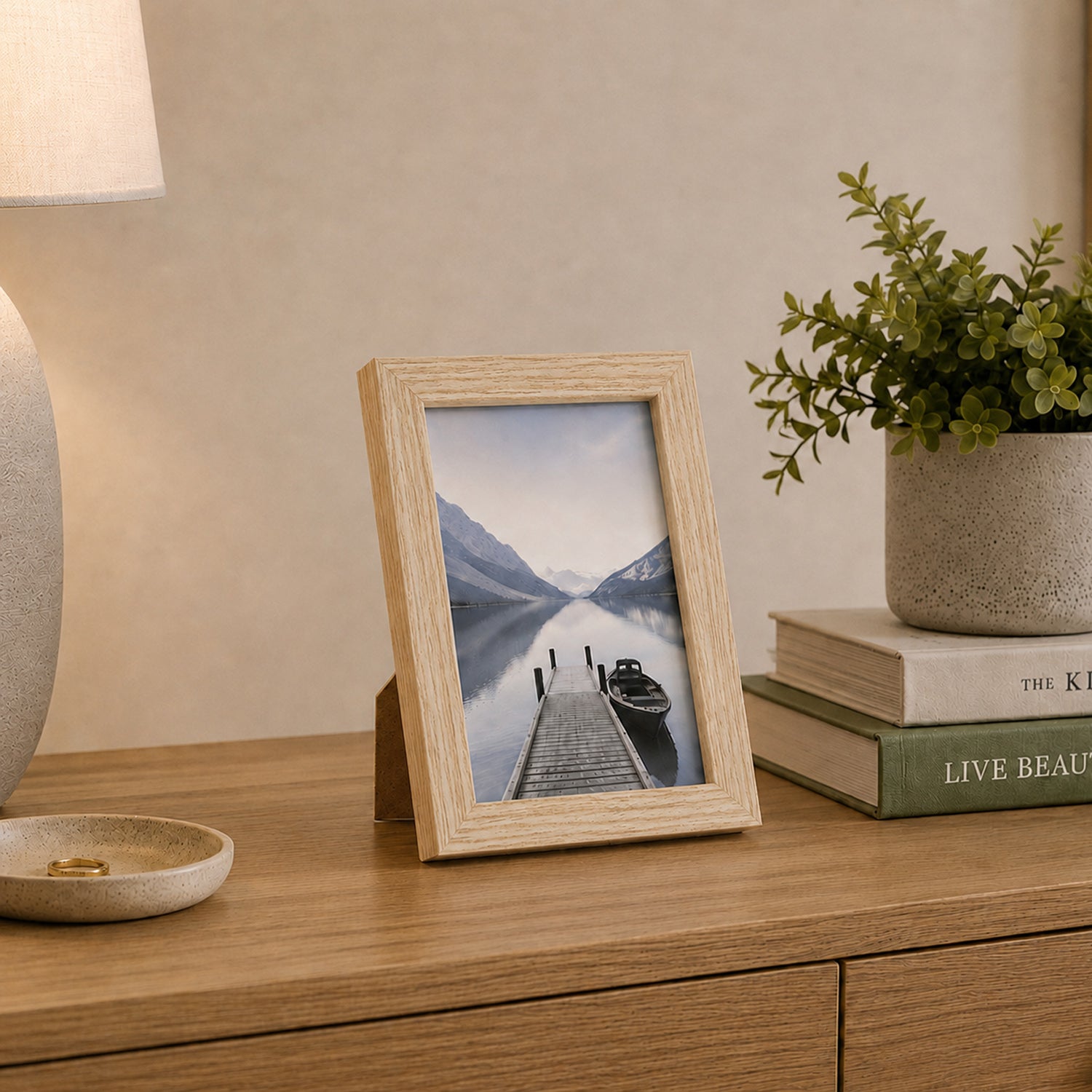 A natural wood-look photo frame displaying a tranquil lakeside landscape is styled on a timber console alongside books, greenery, and a ceramic lamp for a warm, contemporary look.