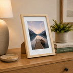 A natural wood-look photo frame displaying a tranquil lakeside landscape is styled on a timber console with books, greenery, and a ceramic lamp for a warm, contemporary look.
