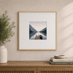 A natural wood-look square photo frame with a wide white mat elegantly displays a tranquil lakeside landscape, styled above a timber sideboard in a warm, contemporary interior.