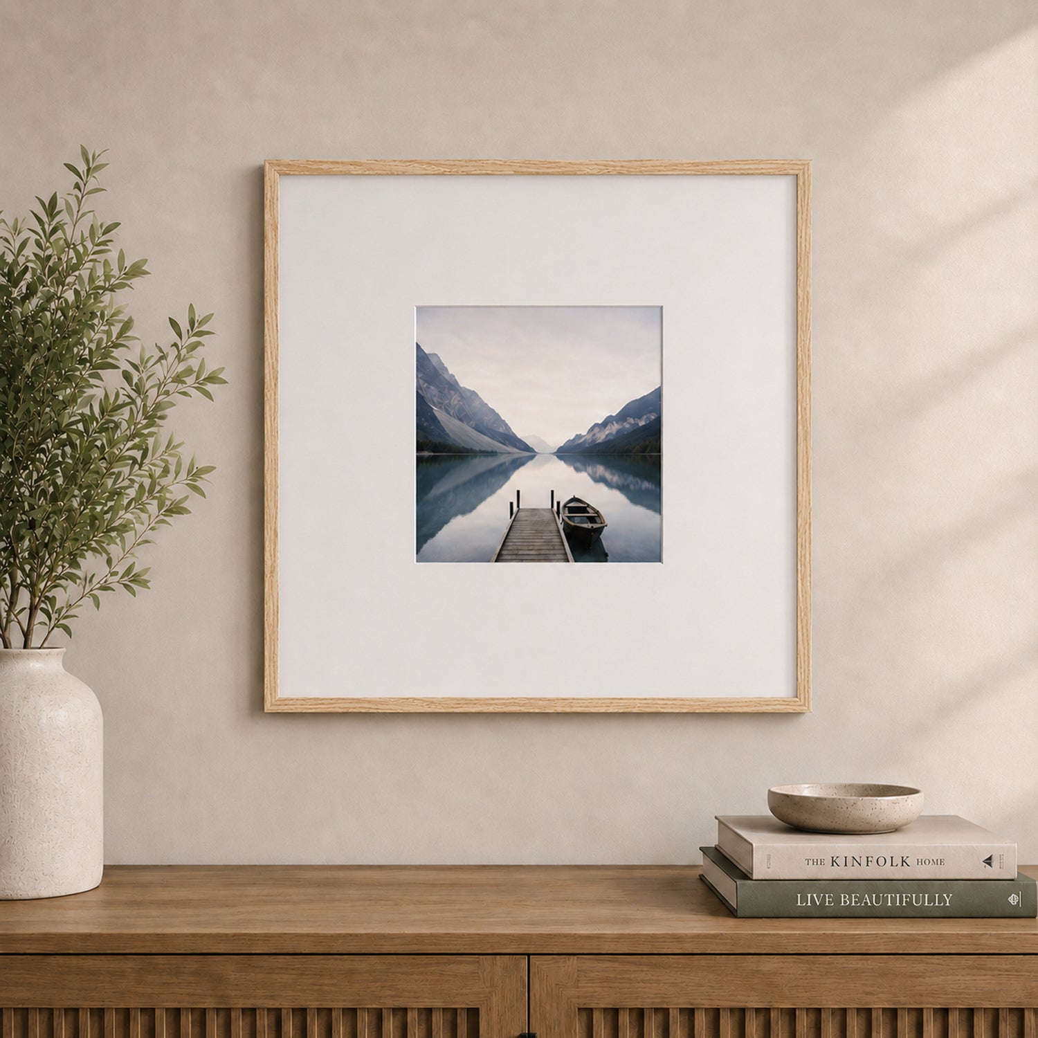 A natural wood-look square photo frame with a wide white mat elegantly displays a tranquil lakeside landscape, styled above a timber sideboard in a warm, contemporary interior.