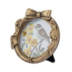 An oval decorative picture frame with a metallic gold finish, featuring a bow at the top and a bird perched among yellow flowers on the insert, shown on a plain white background.