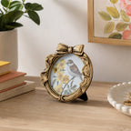 A small oval decorative picture frame with a bow at the top, shown standing on a wooden table in a styled room. The frame has a distressed gold finish and contains a printed bird-and-floral illustration.