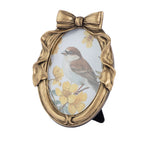 An oval decorative picture frame with a sculpted gold-tone bow at the top and a bird perched among yellow flowers on the insert, shown against a plain white background.