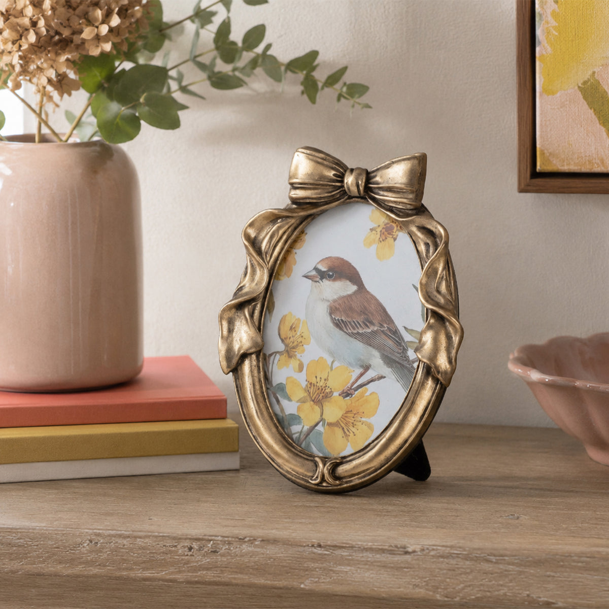 An oval decorative picture frame with a metallic gold bow at the top, displayed on a wooden surface in a styled room setting next to a vase and books.