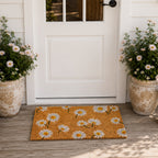A cheerful daisy-print coir doormat adds a fresh, welcoming touch to this bright cottage-inspired entryway, framed by white flowering plants and a classic white front door.