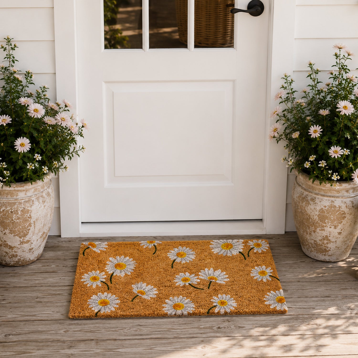 A cheerful daisy-print coir doormat adds a fresh, welcoming touch to this bright cottage-inspired entryway, framed by white flowering plants and a classic white front door.