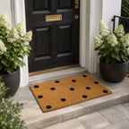 A classic natural coir doormat with a playful black polka dot design adds a stylish, welcoming touch to this elegant modern entryway, framed by lush white hydrangeas and a black front door.