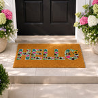 A cheerful "HELLO" coir doormat with colourful floral lettering creates a warm, inviting entrance, beautifully framed by blooming hydrangeas and a classic black front door.