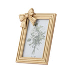 A gold picture frame with a decorative bow on the top left corner, holding a white floral print, shown on a plain white background.