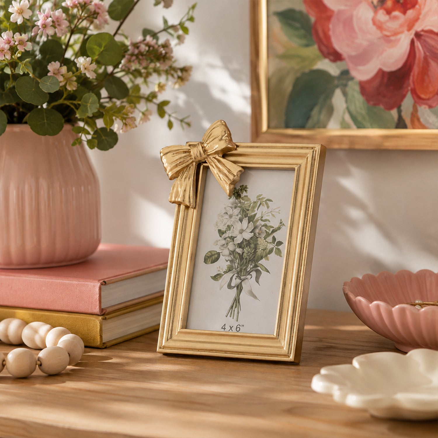 A gold picture frame with a decorative bow on the top left corner, holding a vintage-style floral illustration. The frame is styled on a wooden table next to a pink vase of flowers, books, and decorative bowls.