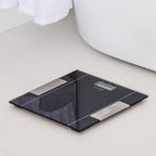 A sleek black digital body scale with a glossy glass surface and silver metallic accents, placed on a light beige tiled bathroom floor next to a white bathtub.
