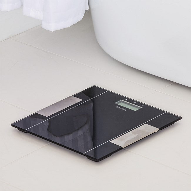 A sleek black digital body scale with a glossy glass surface and silver metallic accents, placed on a light beige tiled bathroom floor next to a white bathtub.