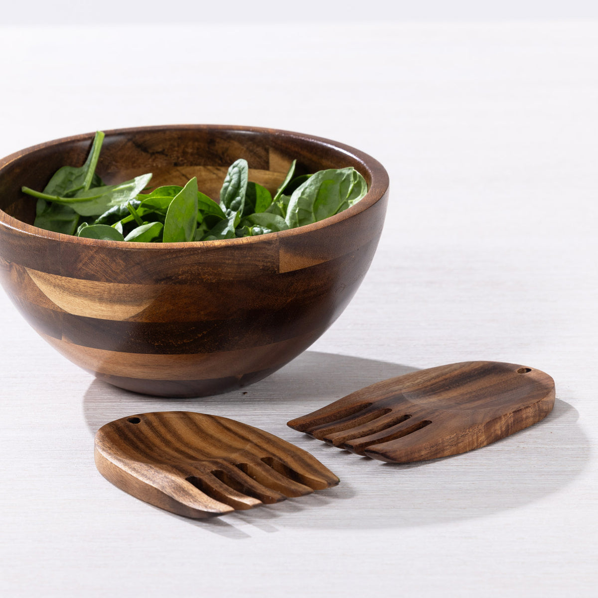 A dark brown wooden salad bowl filled with green spinach leaves, shown with two matching wooden salad servers on a light gray surface.