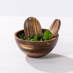 A small wooden salad bowl with two wooden serving utensils, filled with green leafy spinach, on a light gray surface with a white background.