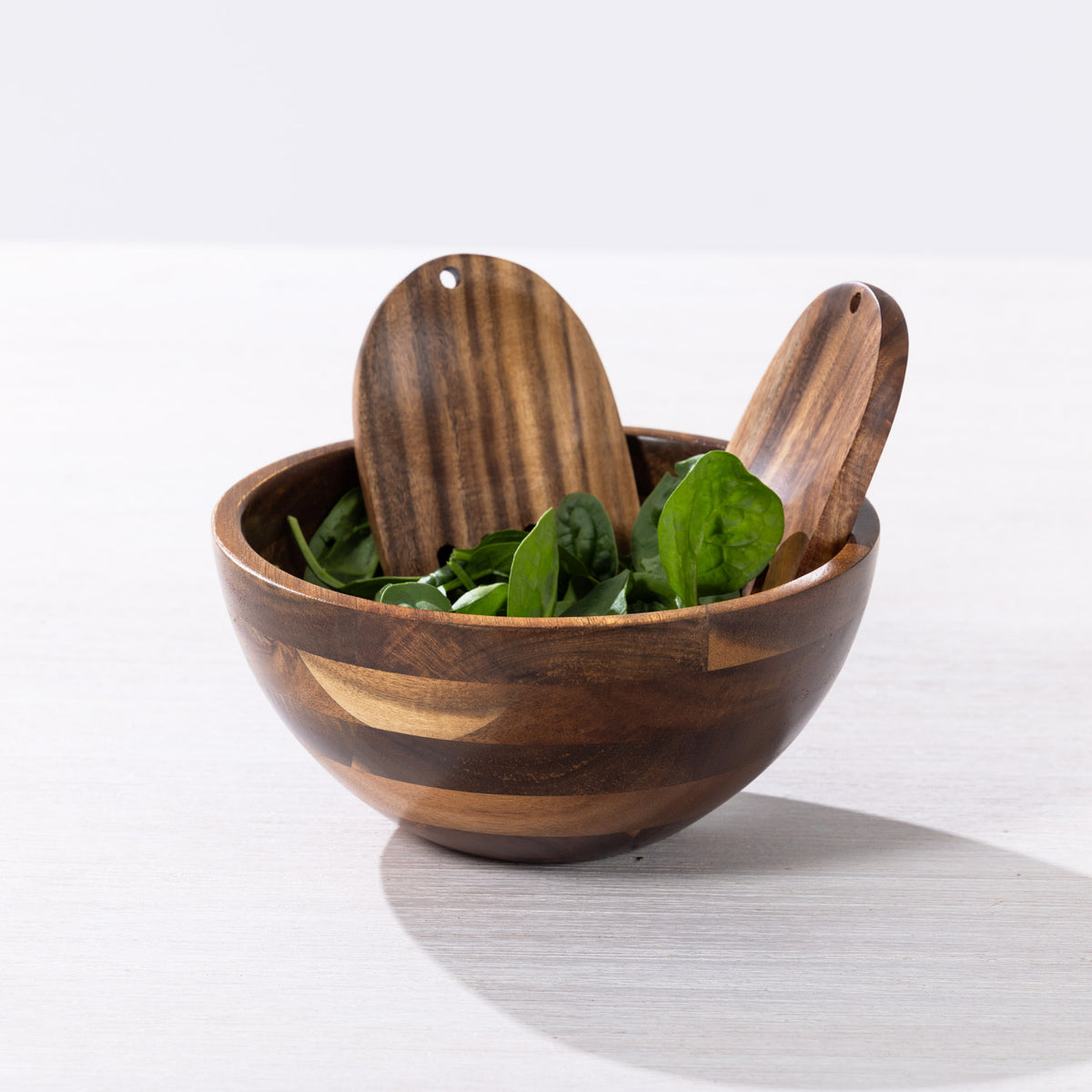 A small wooden salad bowl with two wooden serving utensils, filled with green leafy spinach, on a light gray surface with a white background.