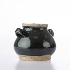 A small handmade ceramic pot with a glossy black glaze and natural beige clay base, featuring two side handles, photographed on a white background.