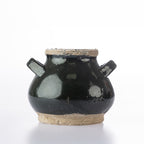 A small handmade ceramic pot with a glossy black glaze, featuring two side handles, a natural clay rim, and an exposed clay base, photographed on a white background.