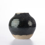 A small round ceramic vase with a glossy black glaze and a natural beige clay foot, photographed on a clean white background.