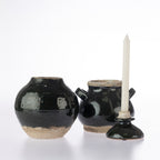 Three rustic black glazed ceramic vessels on a white background, including a round vase, a small pot with side handles, and a candle holder with a white taper candle.
