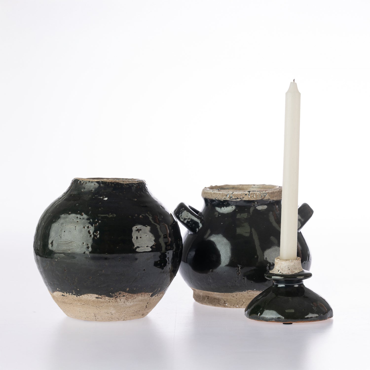Three rustic black glazed ceramic vessels on a white background, including a round vase, a small pot with side handles, and a candle holder with a white taper candle.