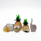 Collection of decorative hand-blown glass ornaments including smoke grey elephants, pineapple figurines, birds, fish, and a swirled glass sphere, displayed against a white background.