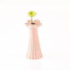 A light pink ceramic vase with a fluted, ribbed body and a flower-shaped rim, holding a single pale yellow-green daisy on a white background.