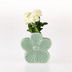 A light green ceramic flower-shaped vase with a ribbed center and five petals, holding white flowers with green stems, set against a plain white background.