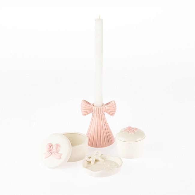 A white taper candle in a pleated pink holder, displayed with two small cream-colored lidded boxes and a small decorative tray, all featuring pink bow details, on a seamless white background.