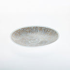 A shallow round glass plate with a textured, sunburst pattern of fine lines in warm brown and white, photographed from directly above on a seamless white background.