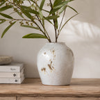 Small reactive glaze ceramic vase displayed with leafy stems on a wooden console table beside stacked books and a decorative bowl in a warm, natural living space.