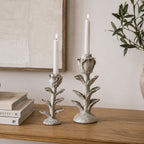 Small and large flower-shaped candle holders displayed with white taper candles on a wooden console table beside stacked books and decorative greenery in a warm, natural living space.