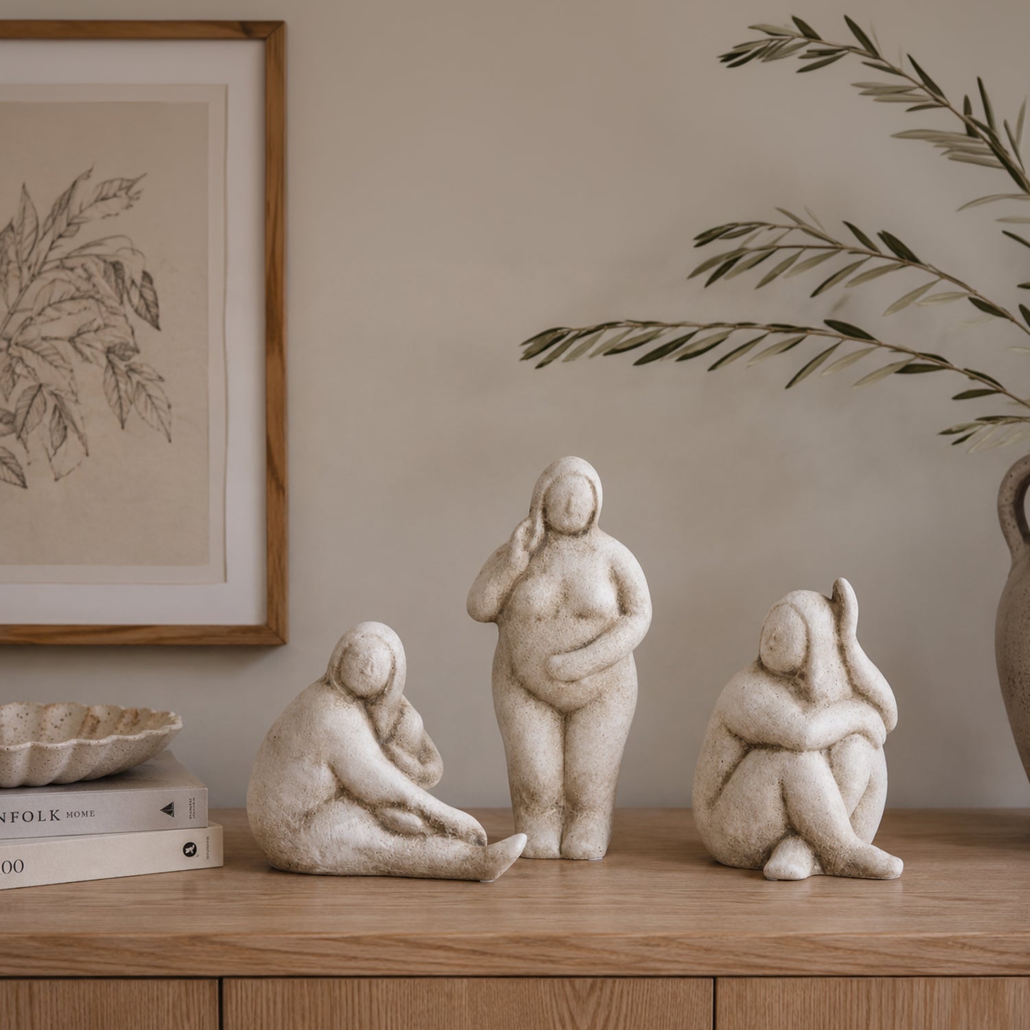 Three natural ceramic lady figurines displayed on a wooden console table with framed botanical artwork, stacked books and olive branches in a warm, neutral interior.