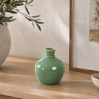 Small glossy green ceramic bud vase styled on a wooden console beside a textured stone-look vase with olive branches and framed artwork in a warm, contemporary interior.