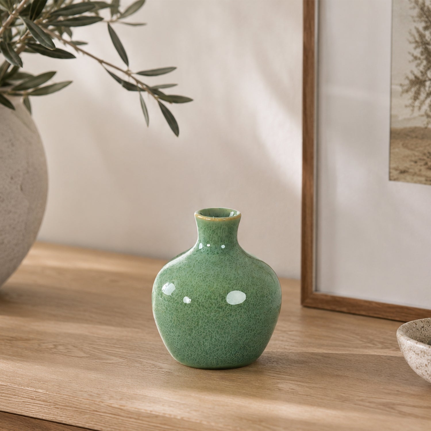 Small glossy green ceramic bud vase styled on a wooden console beside a textured stone-look vase with olive branches and framed artwork in a warm, contemporary interior.