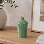 Tall glossy green ceramic vase styled on a wooden console beside a textured stone-look vase with olive branches and framed artwork in a warm, contemporary interior.
