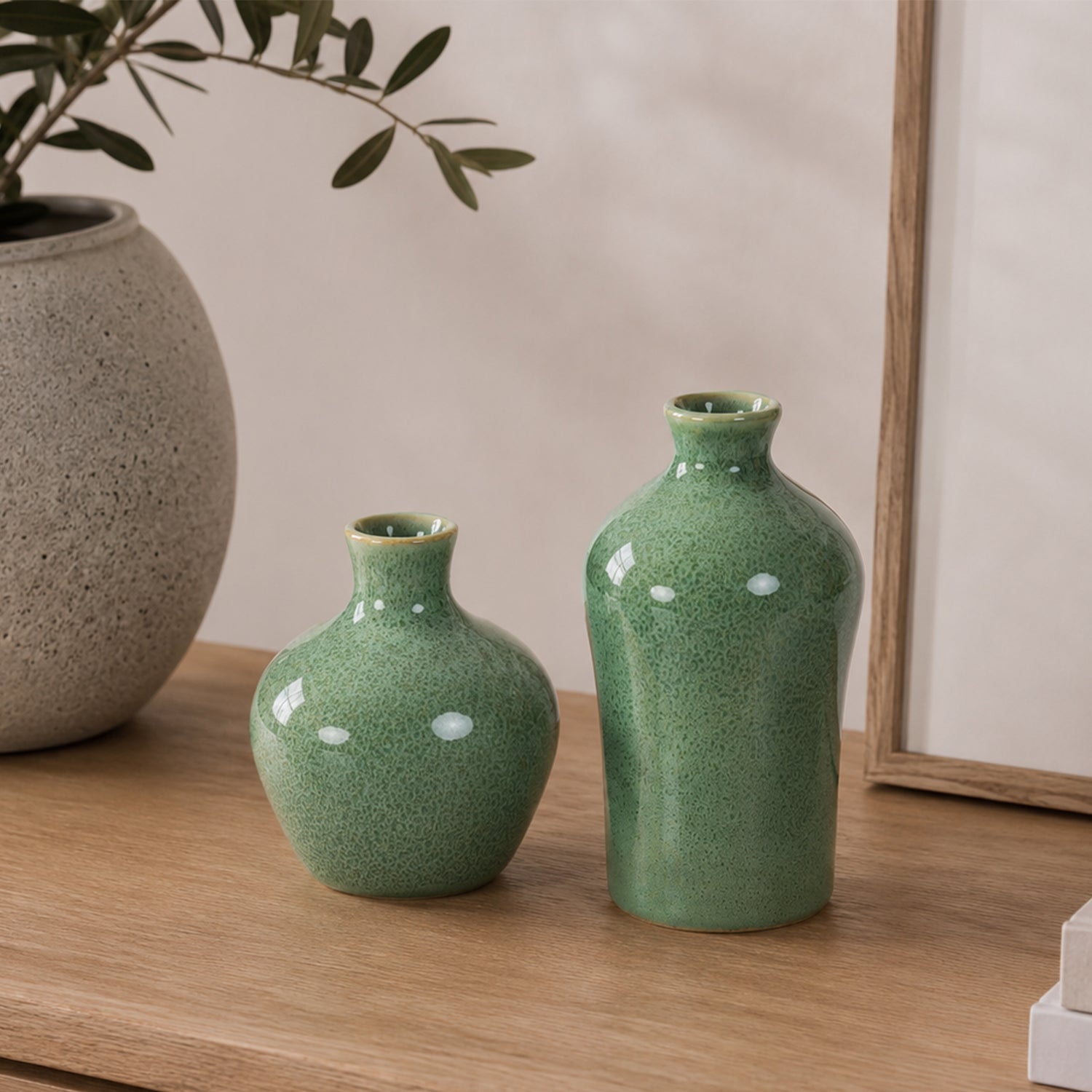 Two green ceramic bud vases displayed on a wooden console beside a textured stone-look vase with olive branches in a warm, contemporary interior.