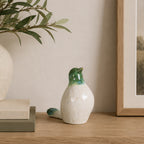 Glossy green and white ceramic bird figurine standing upright on a wooden console beside stacked books, an olive branch arrangement, and framed artwork.