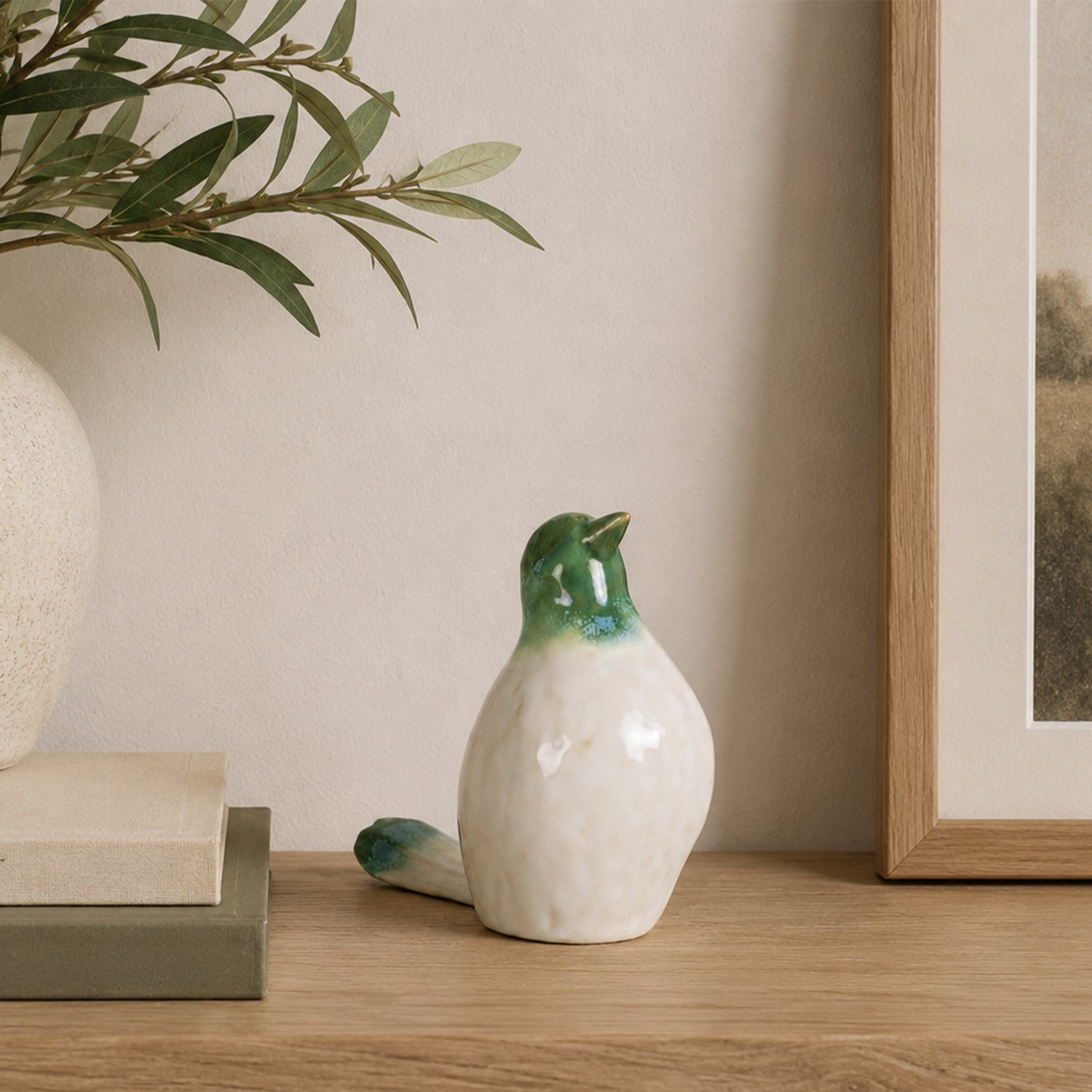 Glossy green and white ceramic bird figurine standing upright on a wooden console beside stacked books, an olive branch arrangement, and framed artwork.