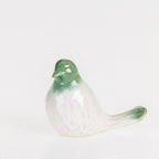 Glossy green and white ceramic bird figurine displayed on a wooden console beside stacked books, an olive branch arrangement, and framed artwork.