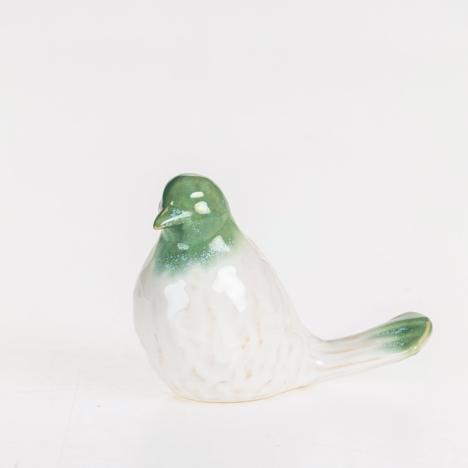 Glossy green and white ceramic bird figurine displayed on a wooden console beside stacked books, an olive branch arrangement, and framed artwork.