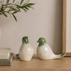 Two green and white ceramic bird figurines styled on a wooden console beside stacked books, an olive branch arrangement, and framed artwork in a warm contemporary interior.