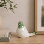 Glossy green and white ceramic bird figurine displayed on a wooden console beside stacked books, an olive branch arrangement, and framed artwork.
