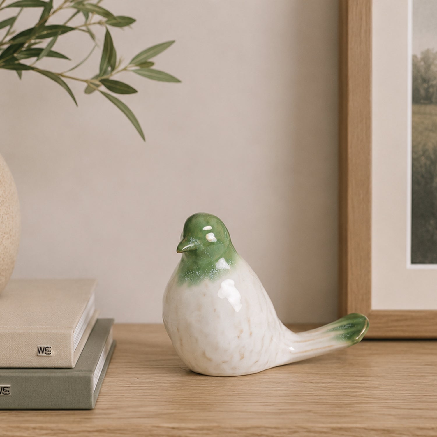 Glossy green and white ceramic bird figurine displayed on a wooden console beside stacked books, an olive branch arrangement, and framed artwork.